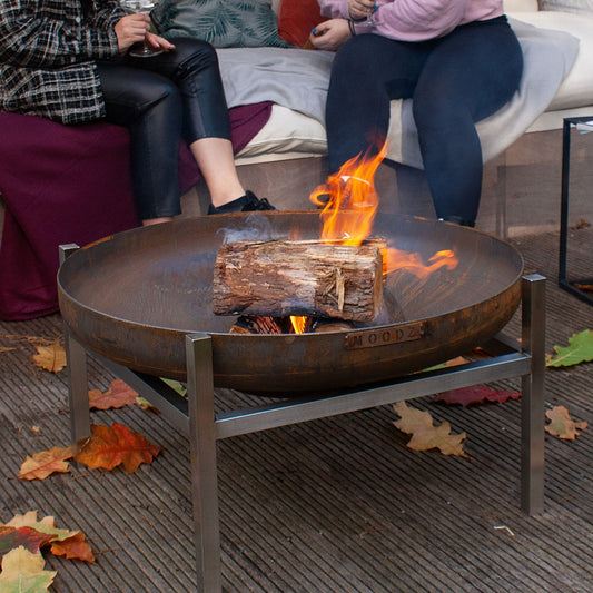 Friends gathered around a lit Moodz Corten steel fire bowl on a wooden deck at dusk, autumn leaves scattered