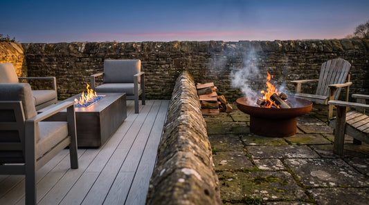 Split dusk garden scene comparing a gas fire pit table with clean blue flame and a wood-burning corten fire pit with glowing logs