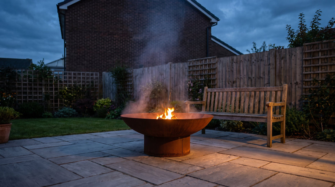 A corten steel fire pit burning on stone paving in a small British back garden at dusk, with a neighbouring house and fence behind