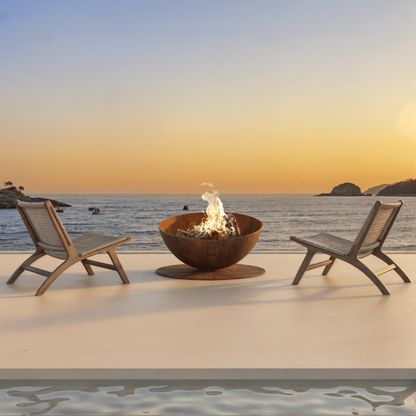 Two lounge chairs by a fire pit on a patio with a scenic ocean view at sunset.