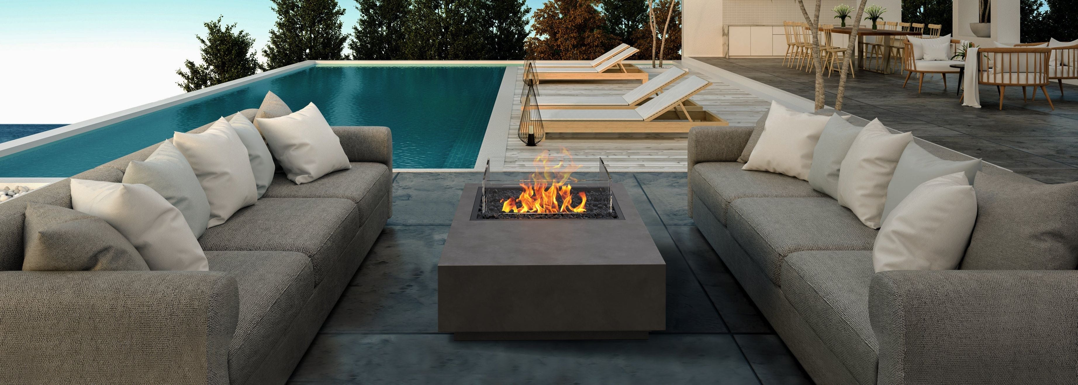 EcoSmart Fire Manhattan 50 gas fire pit table alight beside an infinity pool at dusk, on a covered terrace with grey sectional sofas